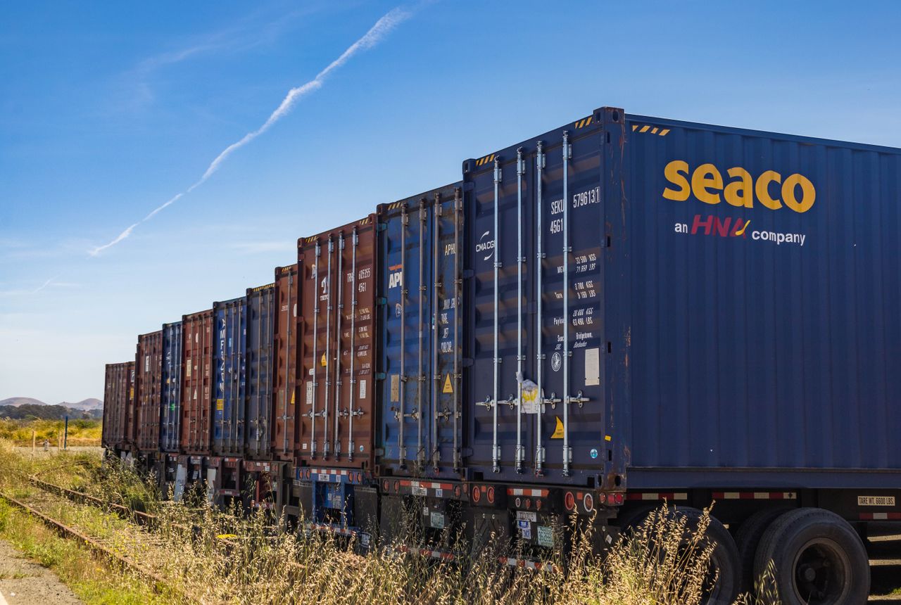 Row of vibrant cargo containers on train tracks under a clear blue sky.